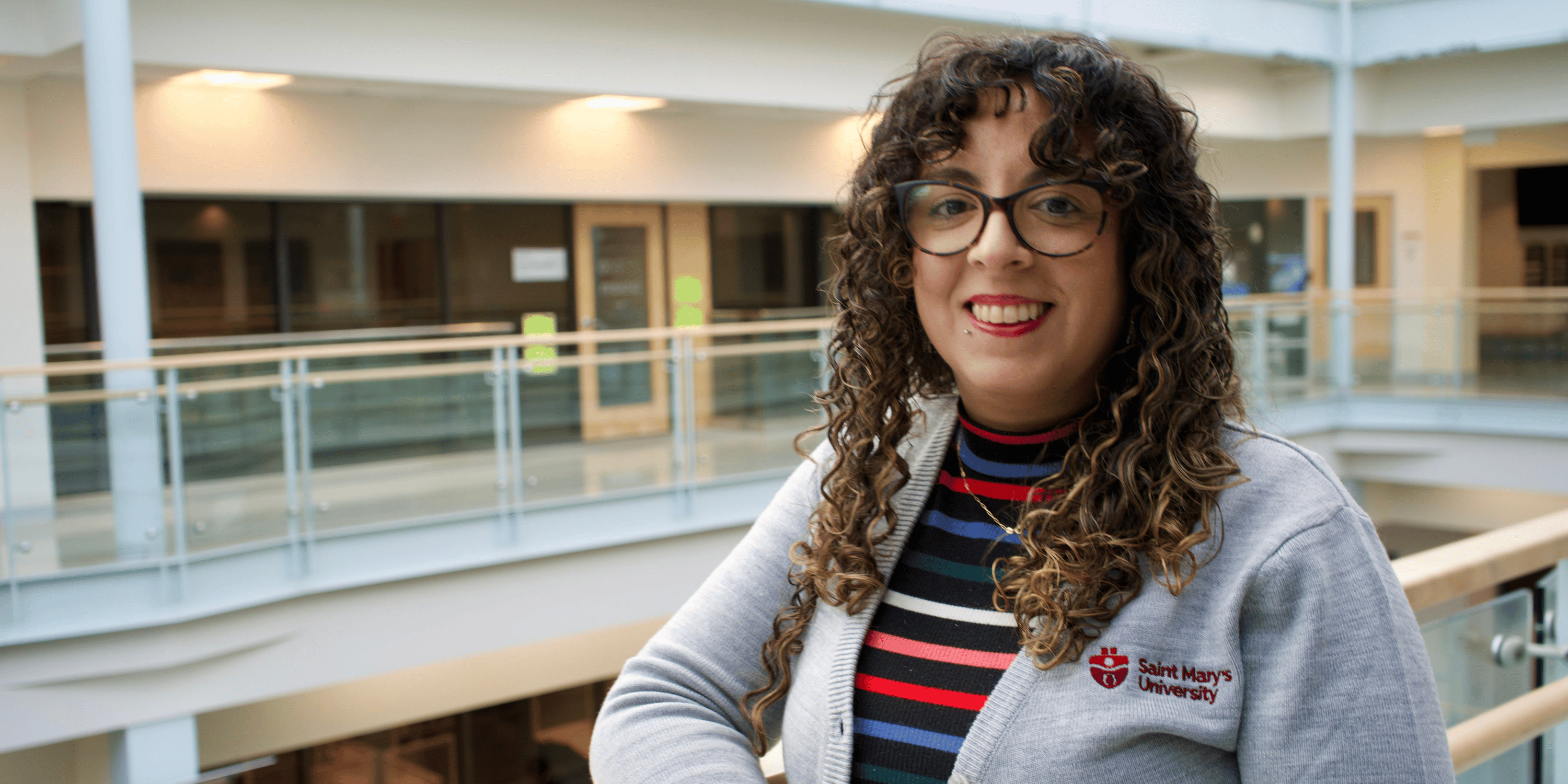 Maria has curly brown hair and glasses. She wears a colourful shirt and Saint Mary's cardigan